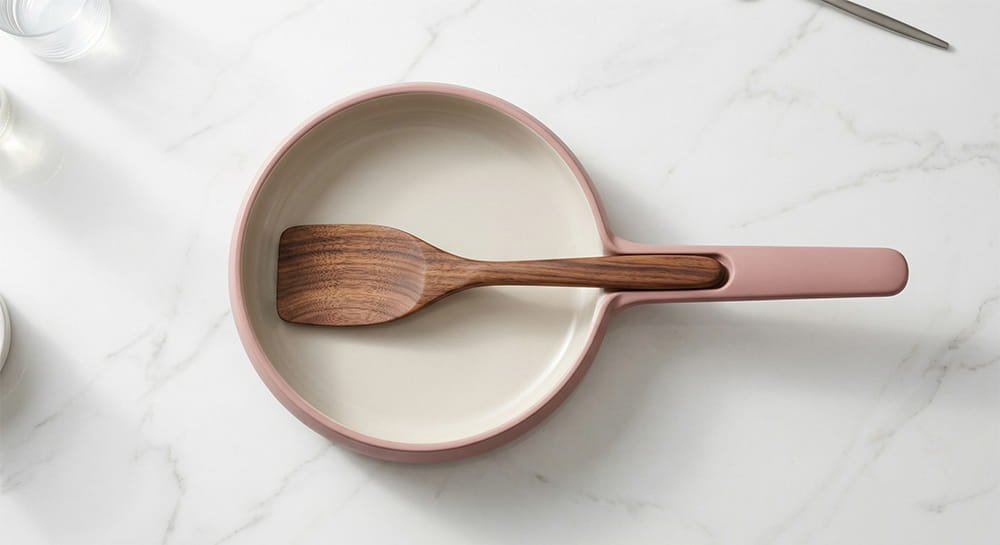 Overhead view of a dusty pink ceramic pan with a custom wooden spatula resting neatly on its built-in handle rest, placed on a white marble counter.